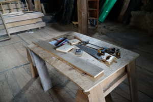 An image of miscellaneous tools used for woodworking placed on a table. Tools include: a hammer, a saw, a ruler, a pencil, a tape measure, and a clamp.