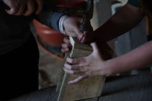 An image of two people building a small wooden birdhouse. The person on the left is holding a nail steady while hammering it. The person on the right is holding the wood in place.