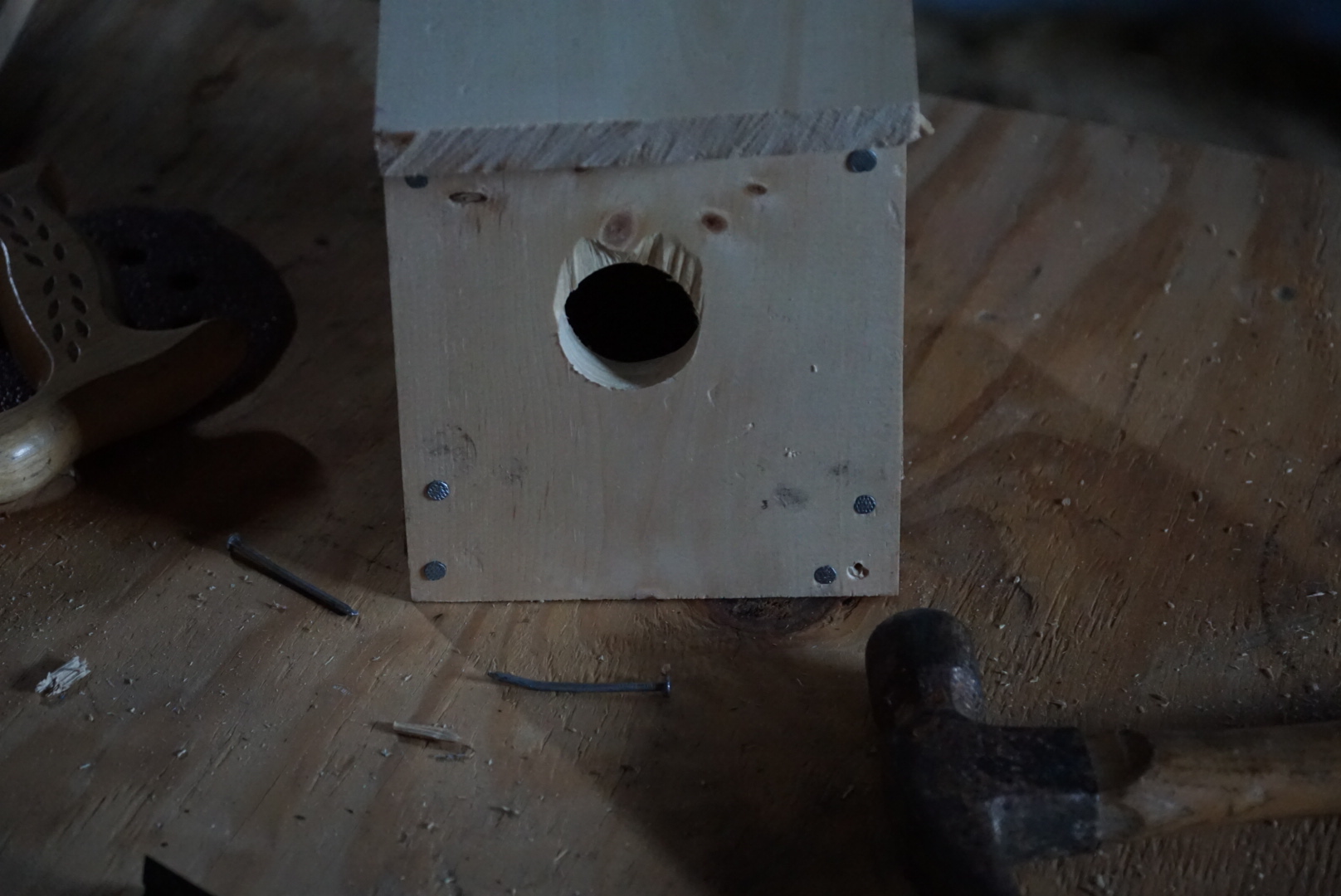 An image of a wooden birdhouse that is sitting on a wooden table. The table also has a saw, building nails and a hammer on it.