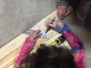 A student uses a drafting triangle and tape measure to mark wood for cutting.