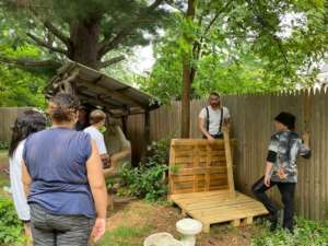 A group of three people stand outdoors watching an instructor show how to assemble a structure using pallets